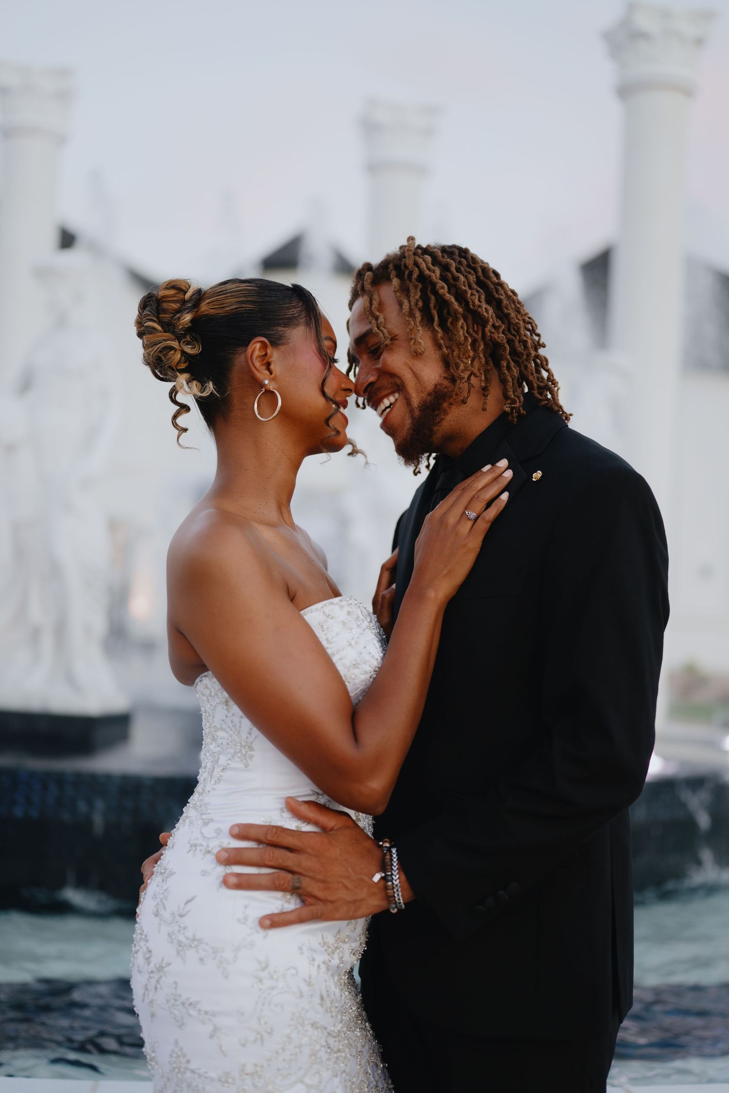 Bride in a lace strapless gown and groom in a black suit touch foreheads beside a fountain with white columns behind.