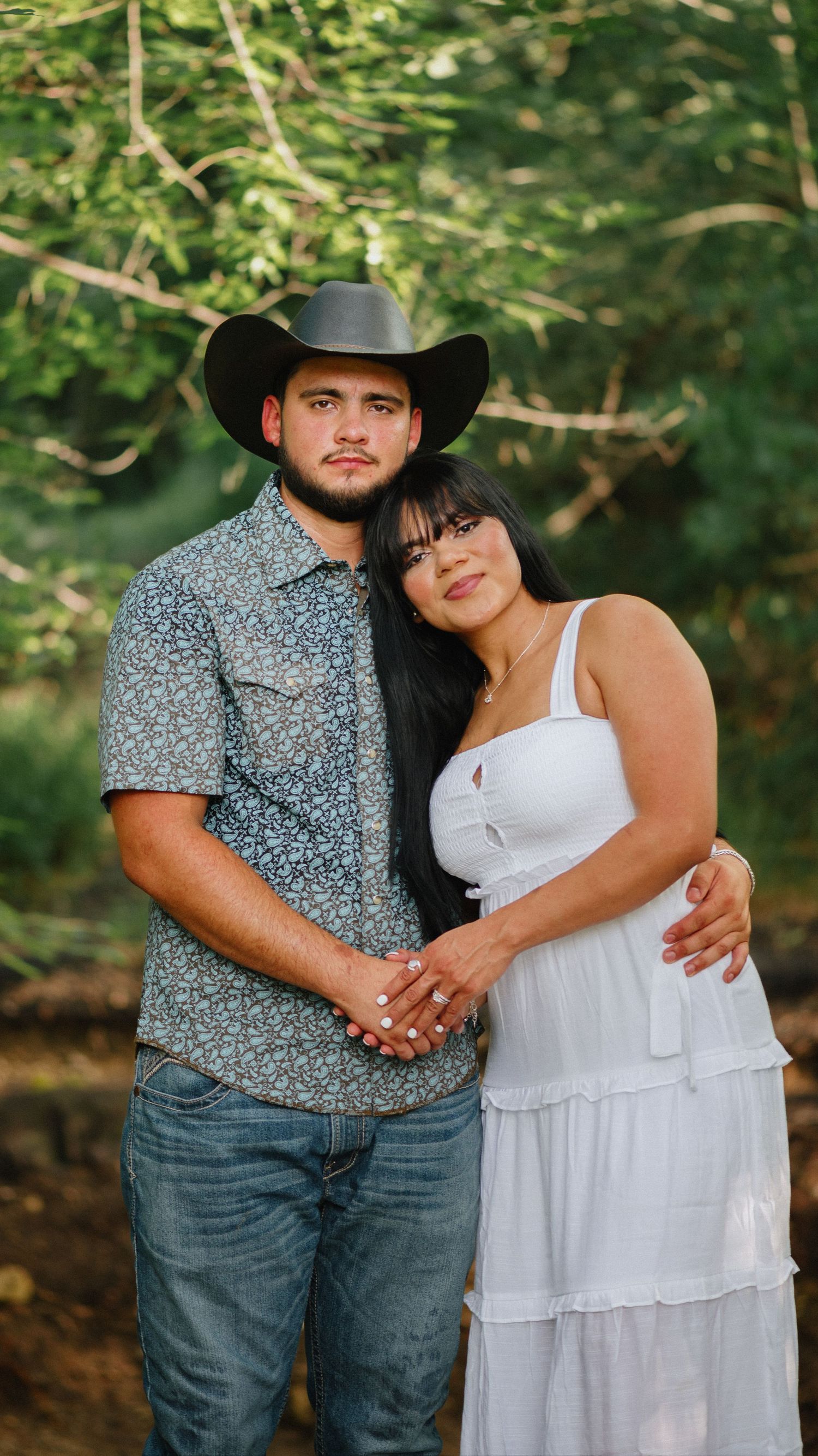Couple embracing outdoors among green trees, a man in a black cowboy hat and patterned shirt beside a woman in a white sundress.