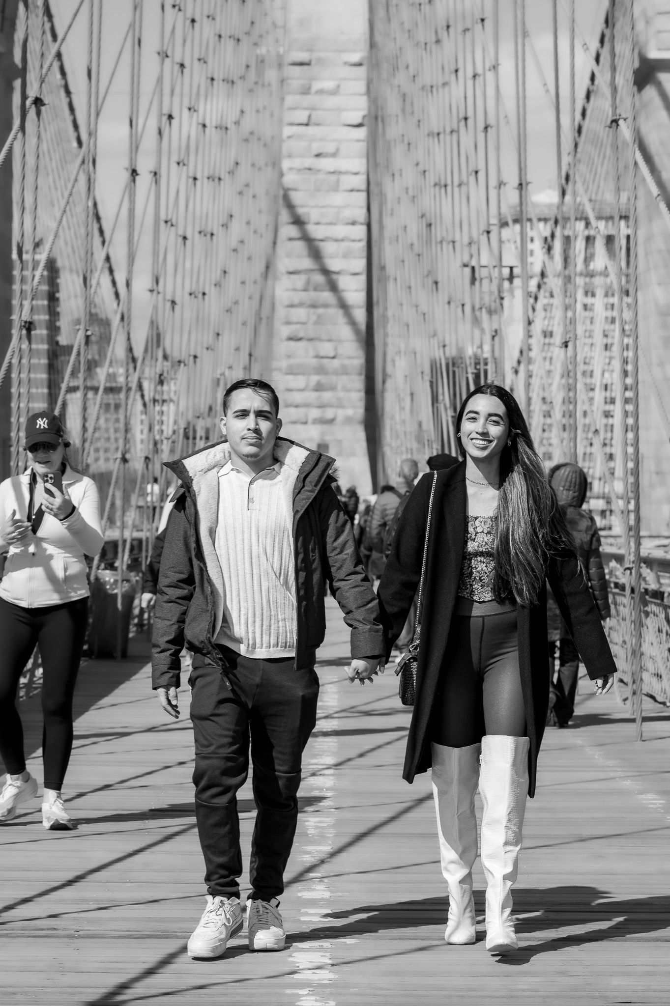 Black and white photo of a smiling couple holding hands and walking across the Brooklyn Bridge with suspension cables behind them.