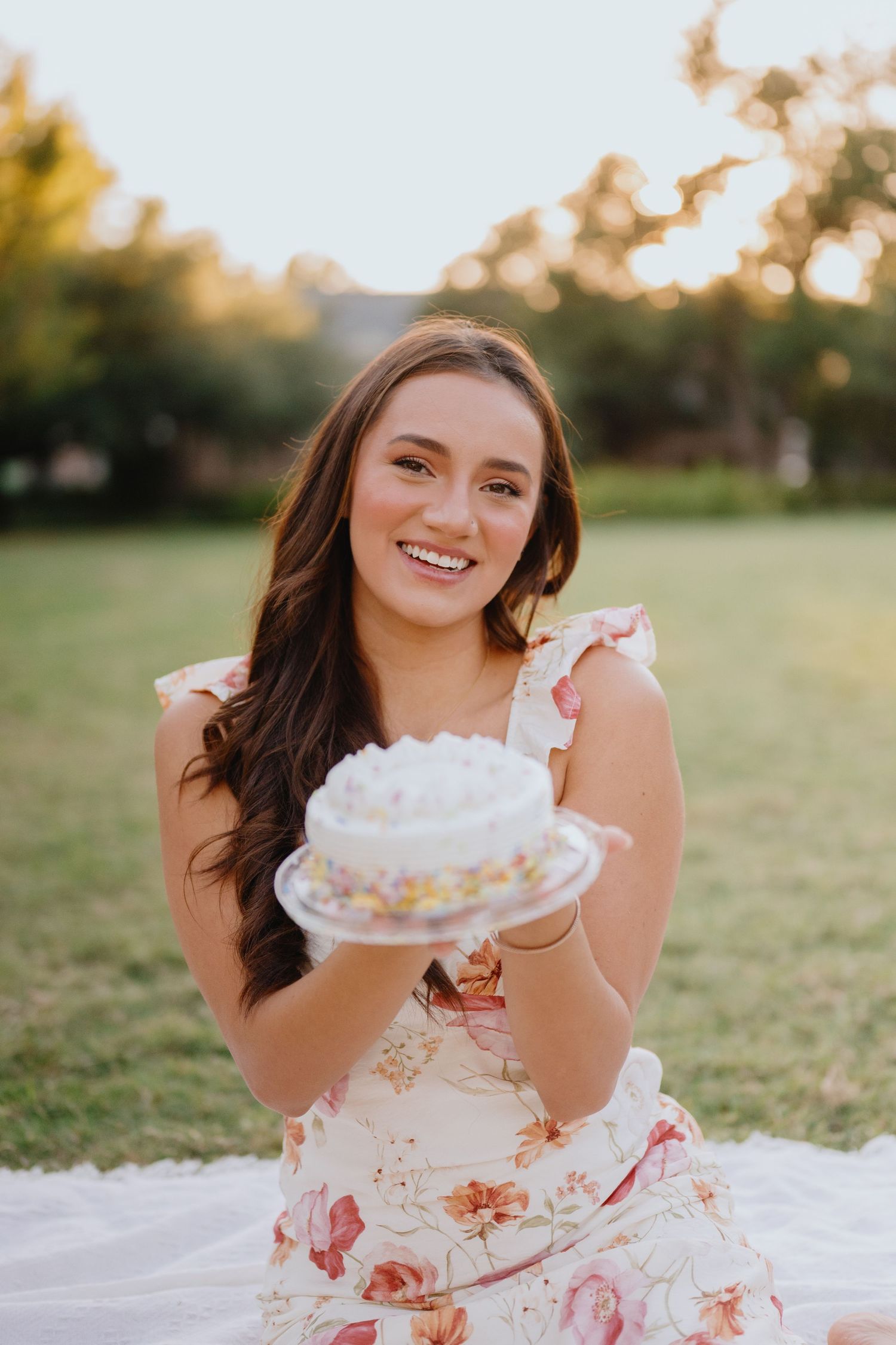 Smiling young woman in a floral dress sitting on a picnic blanket in a park holding a small decorated birthday cake.