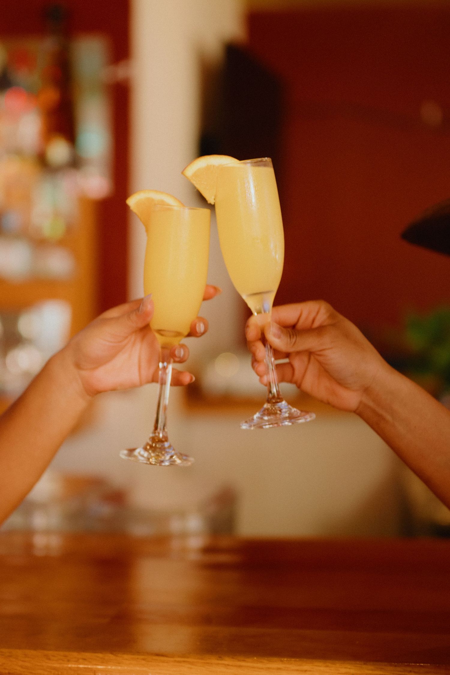 Two hands toast with champagne flutes filled with orange mimosas garnished with orange slices at a wooden bar.