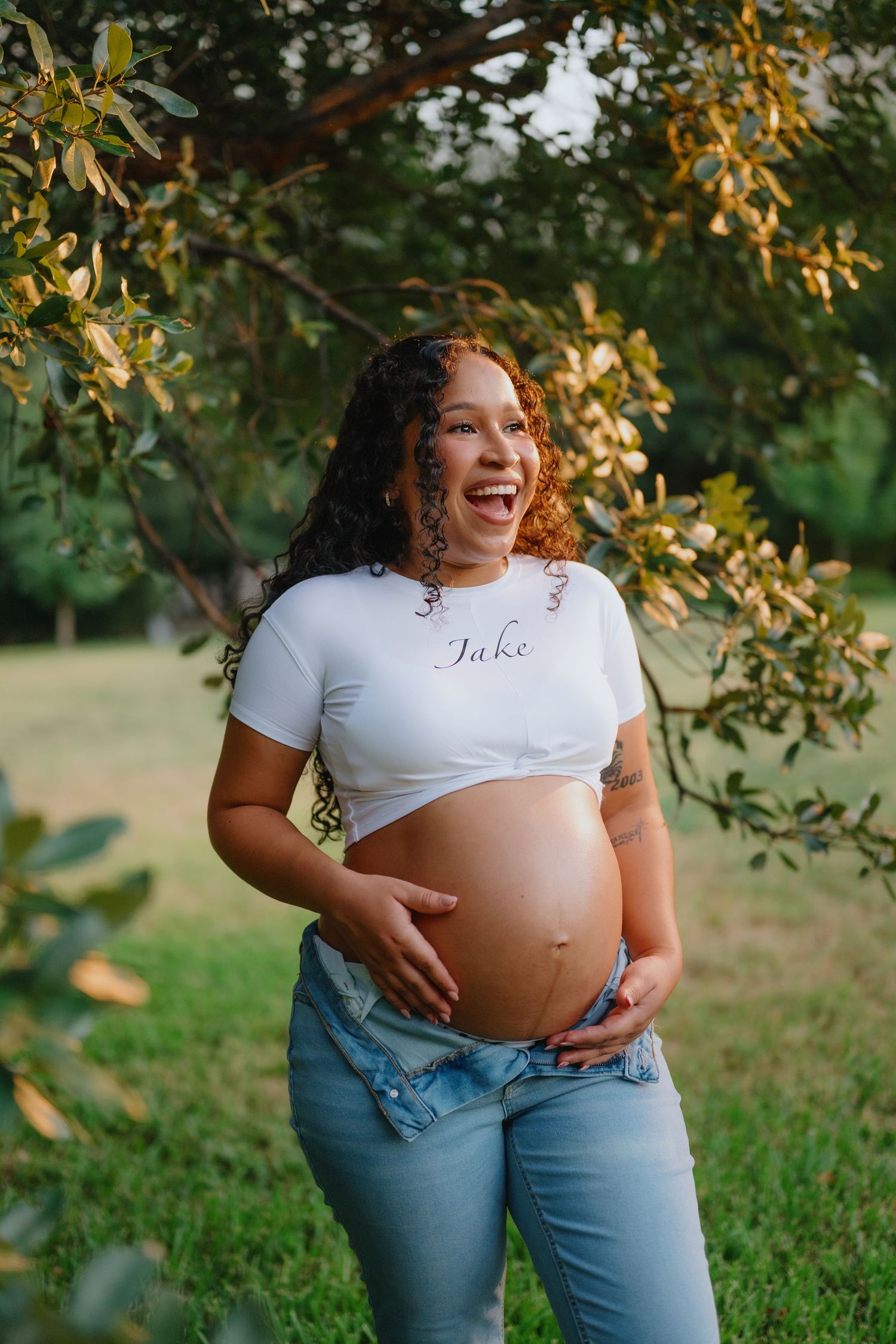 Laughing pregnant woman with curly hair in a white crop top and jeans holds her belly under leafy branches outdoors.