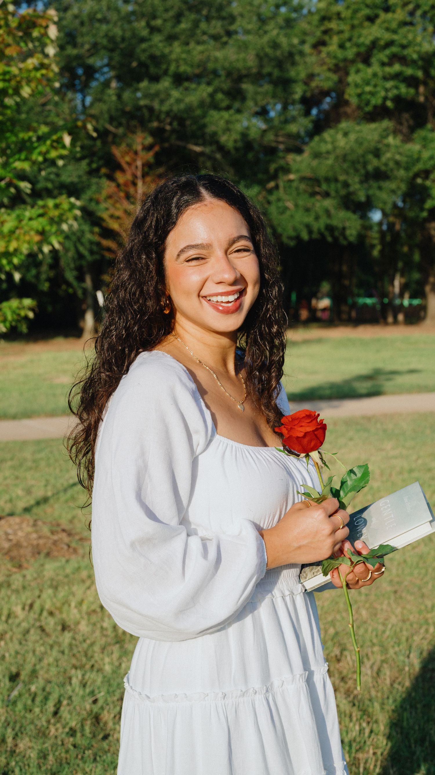 A smiling woman with long curly hair in a white dress holds a red rose and a book in a sunny park.