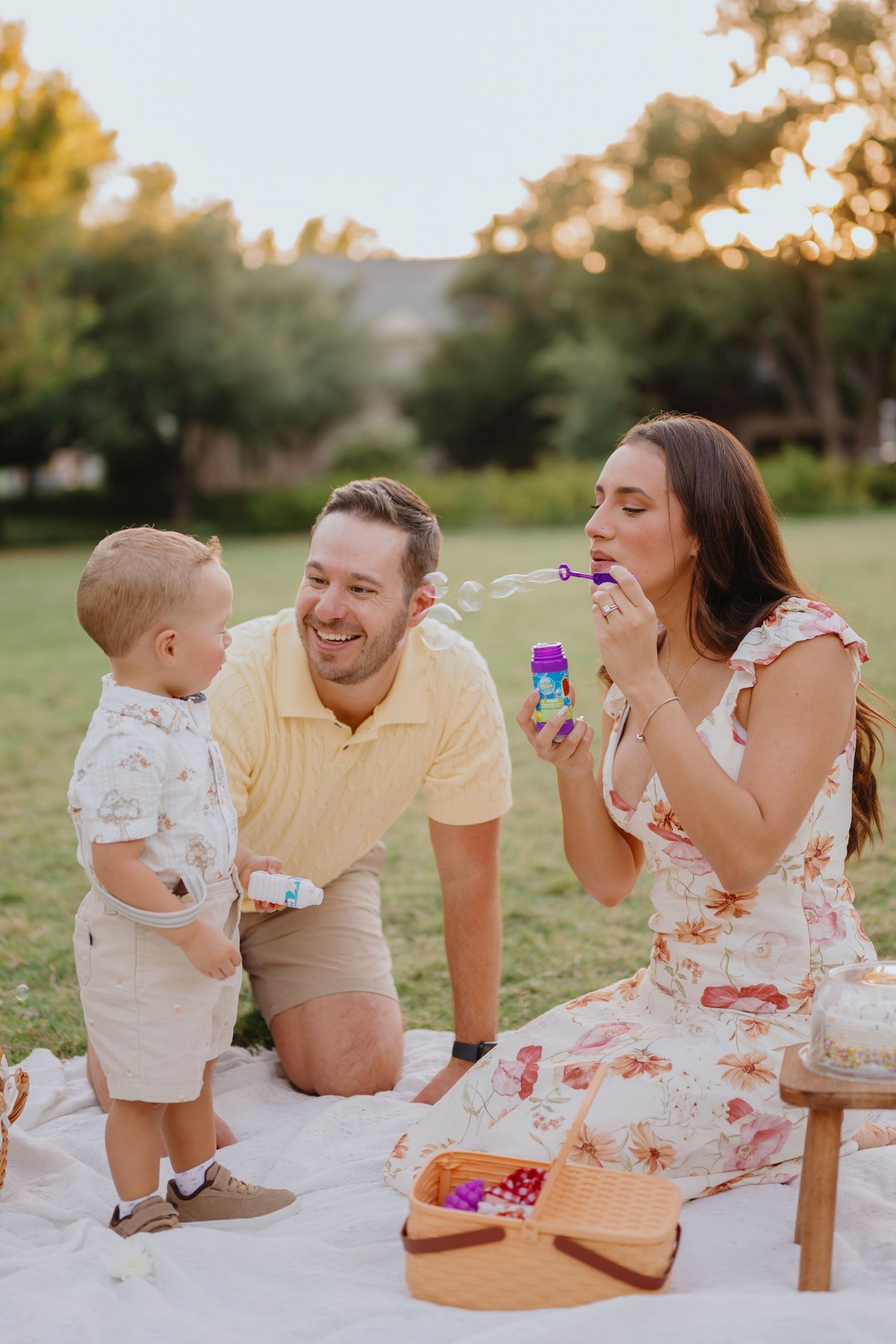 Family enjoys a picnic on a blanket as mother blows bubbles while dad and toddler son smile at sunset.
