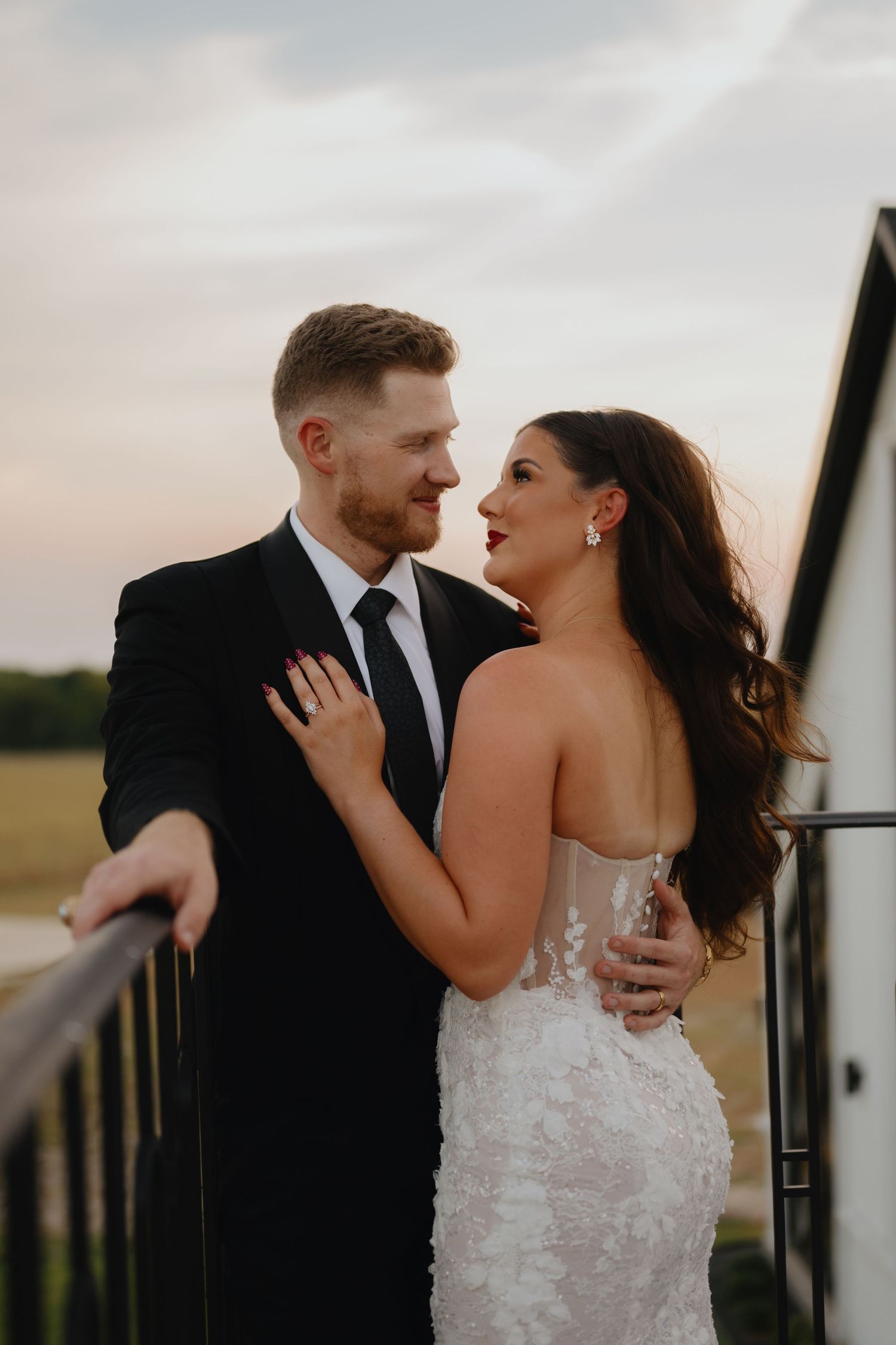 Bride in a floral lace wedding gown and groom in a black suit gaze at each other on a balcony at sunset.