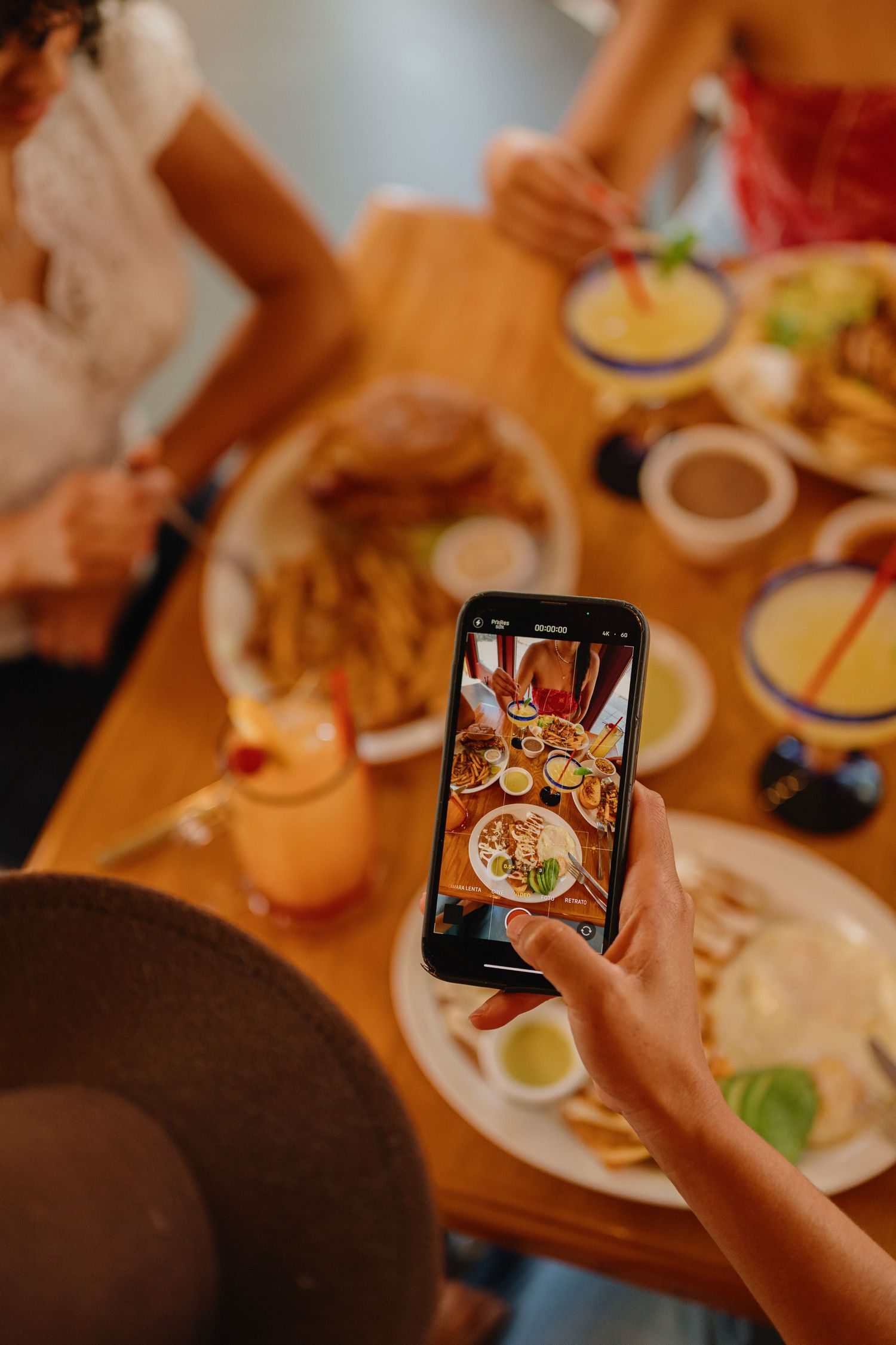 Person photographing a restaurant table filled with burgers, fries, sauces and colorful drinks using a smartphone.