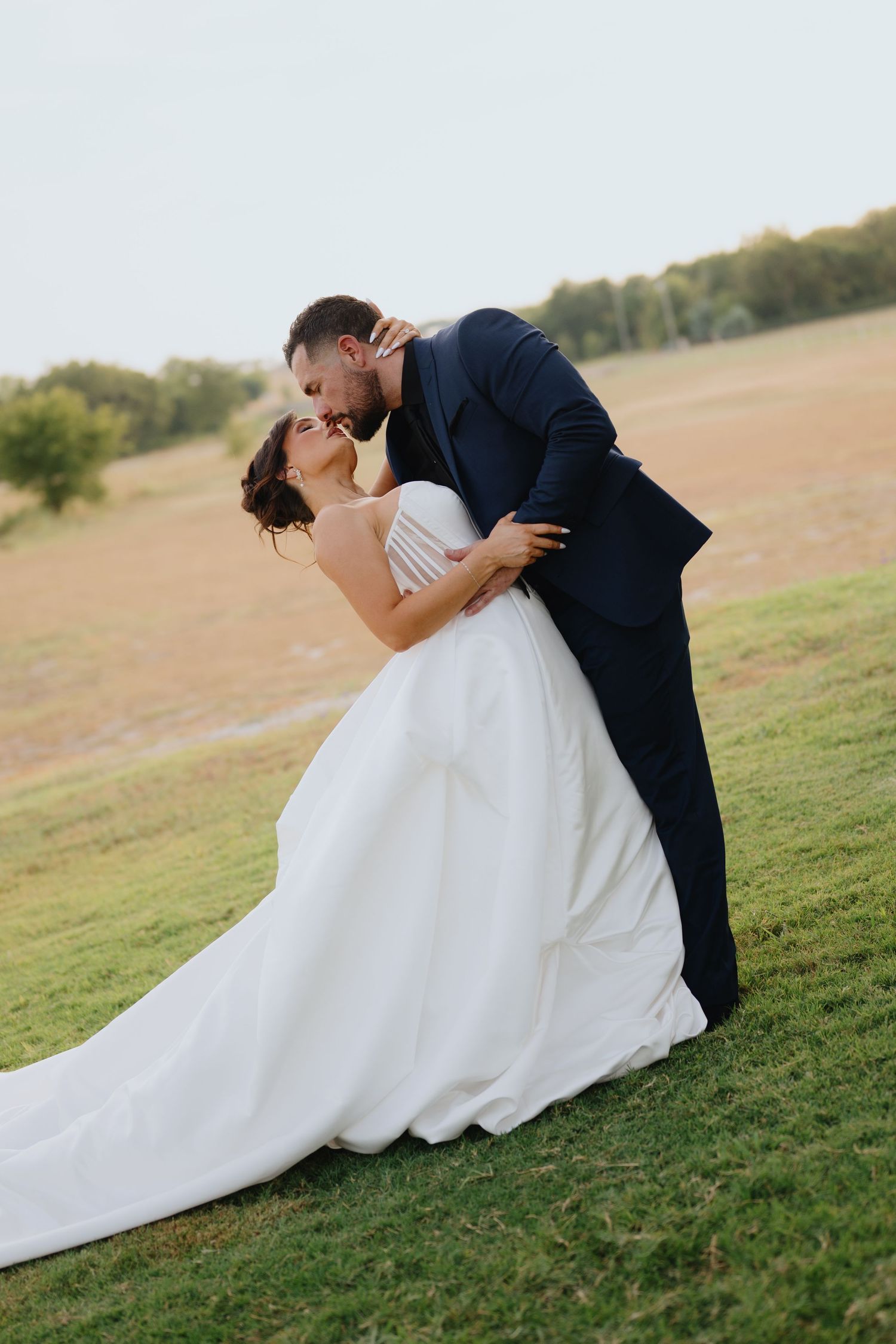 A groom in a navy suit dips his bride in a flowing white gown for a romantic kiss on a grassy hillside at sunset.
