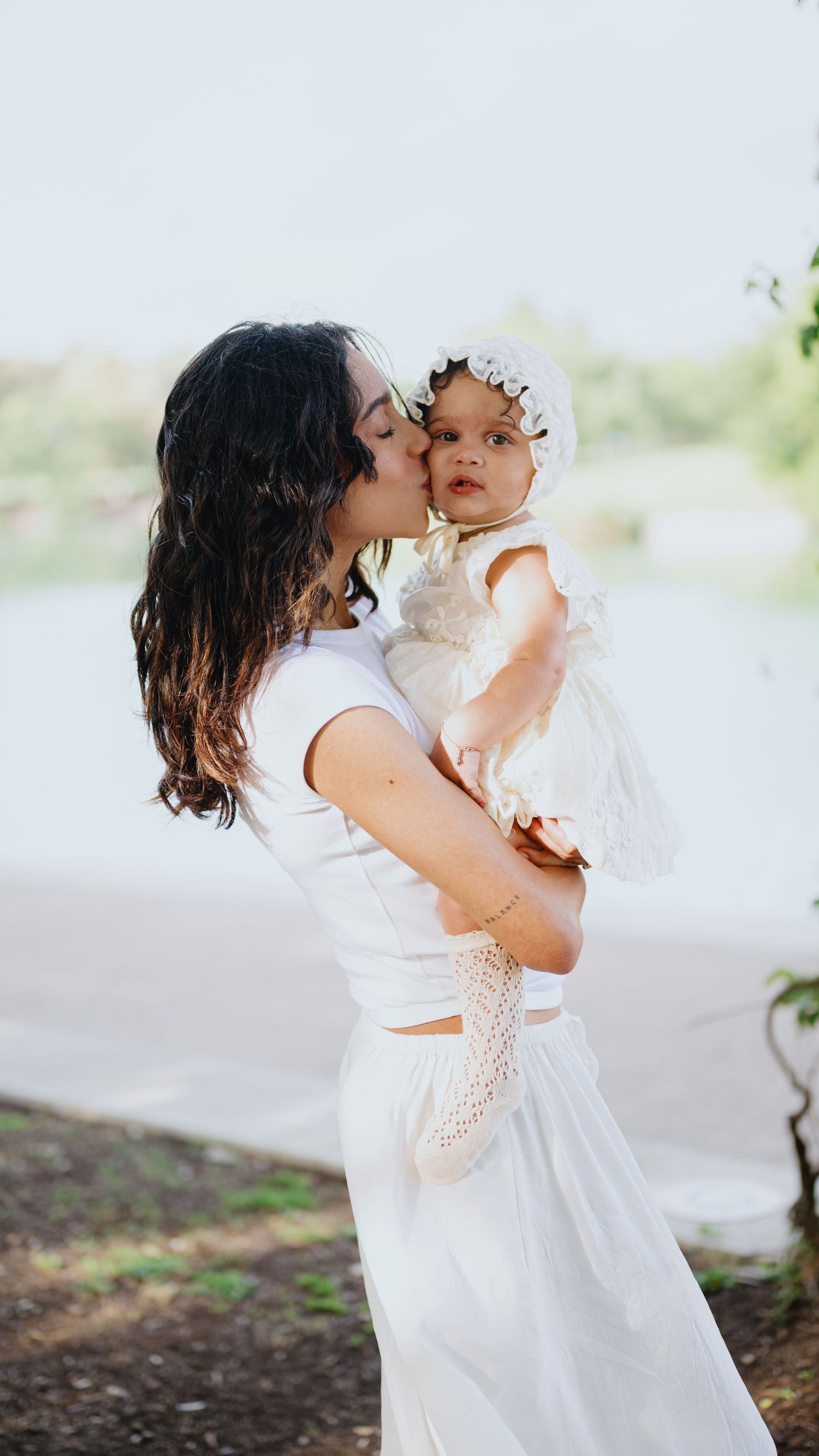 Mother in white kisses her baby girl wearing a lace bonnet and dress during a lakeside family photo session.