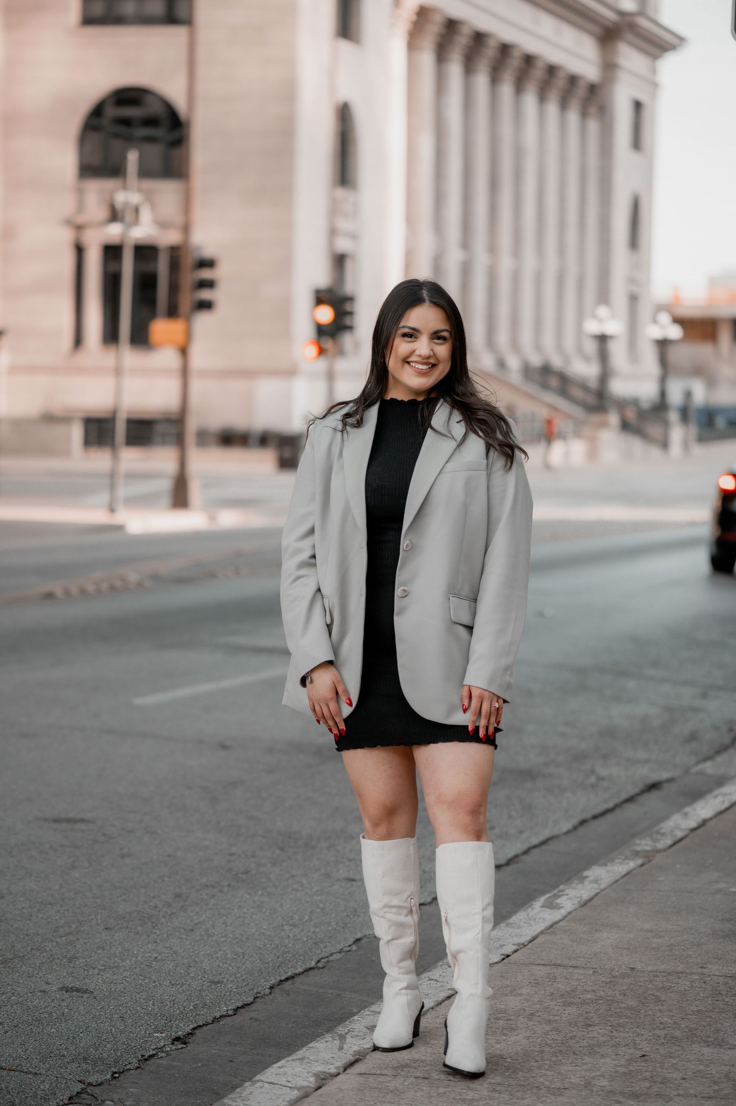 Smiling woman in a gray blazer, black dress and white knee-high boots stands on a city sidewalk near a historic building.