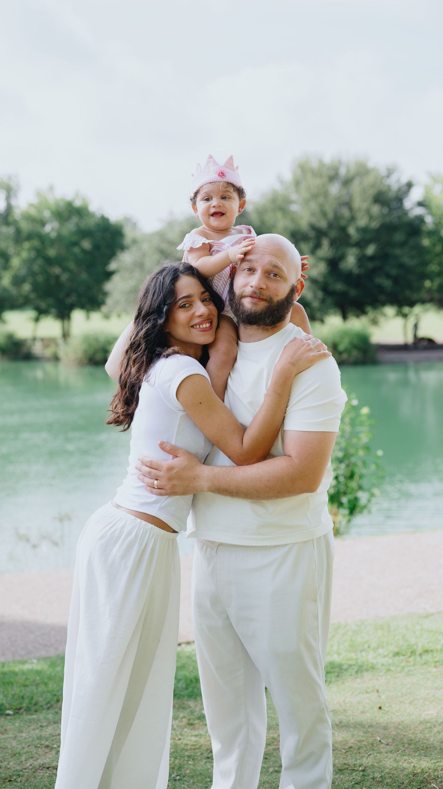 A smiling couple in matching white outfits embrace beside a lake while holding their baby girl wearing a small pink crown.