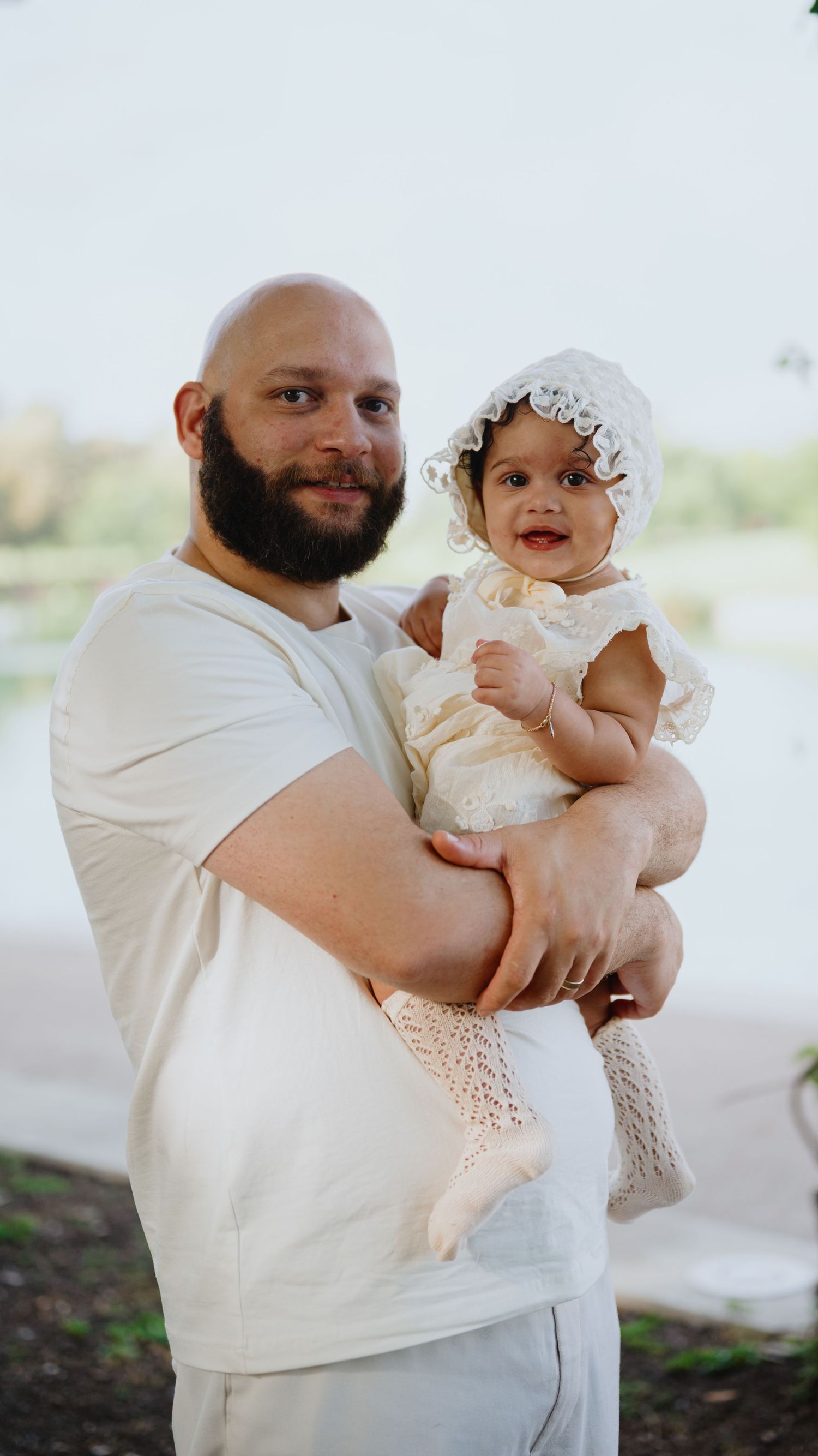 A bearded father in a white t-shirt smiles while holding his baby daughter dressed in a cream lace outfit and bonnet outdoors.