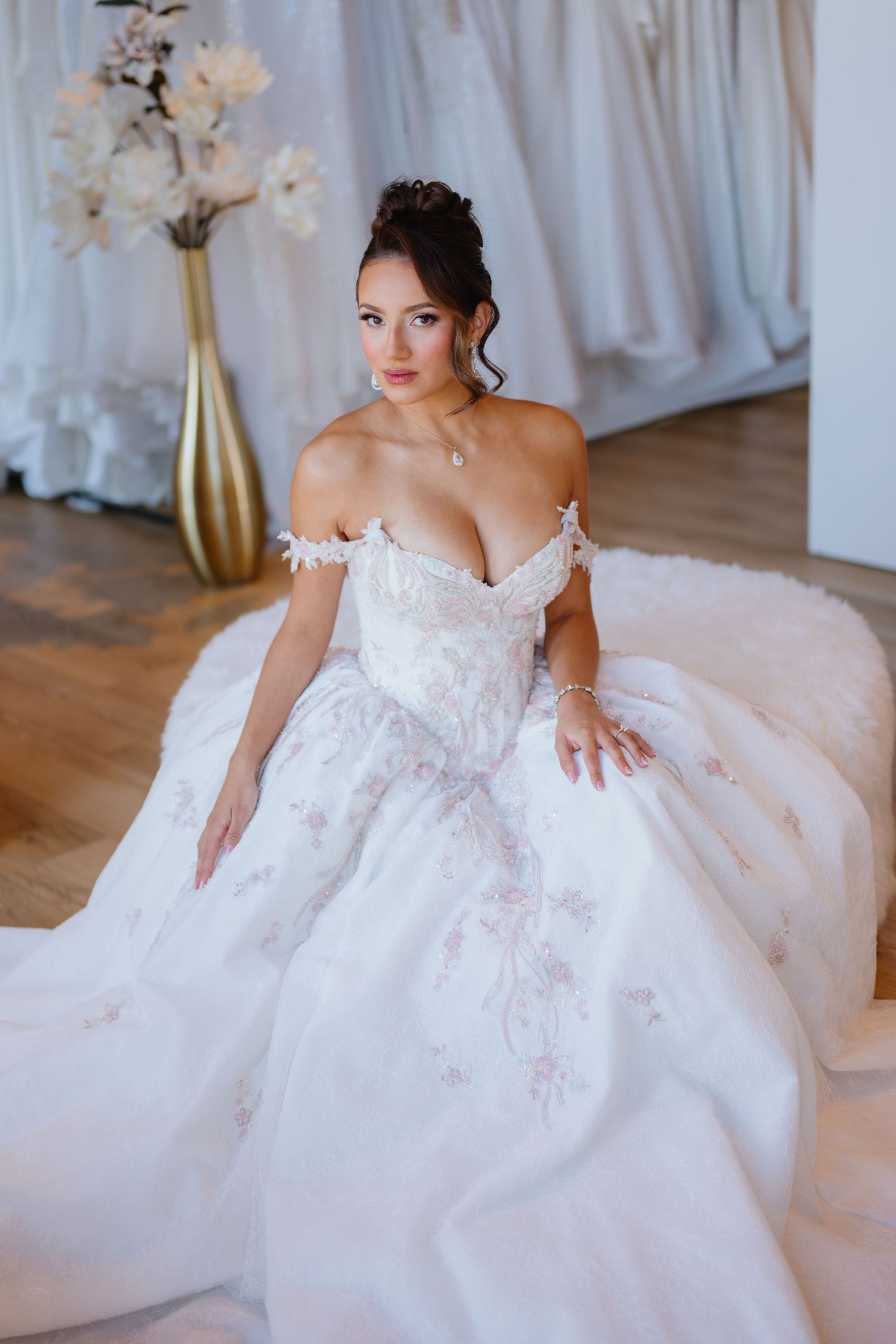 A bride in an off-the-shoulder white ball gown with floral embroidery sits gracefully in a bridal boutique beside a gold vase of blossoms.