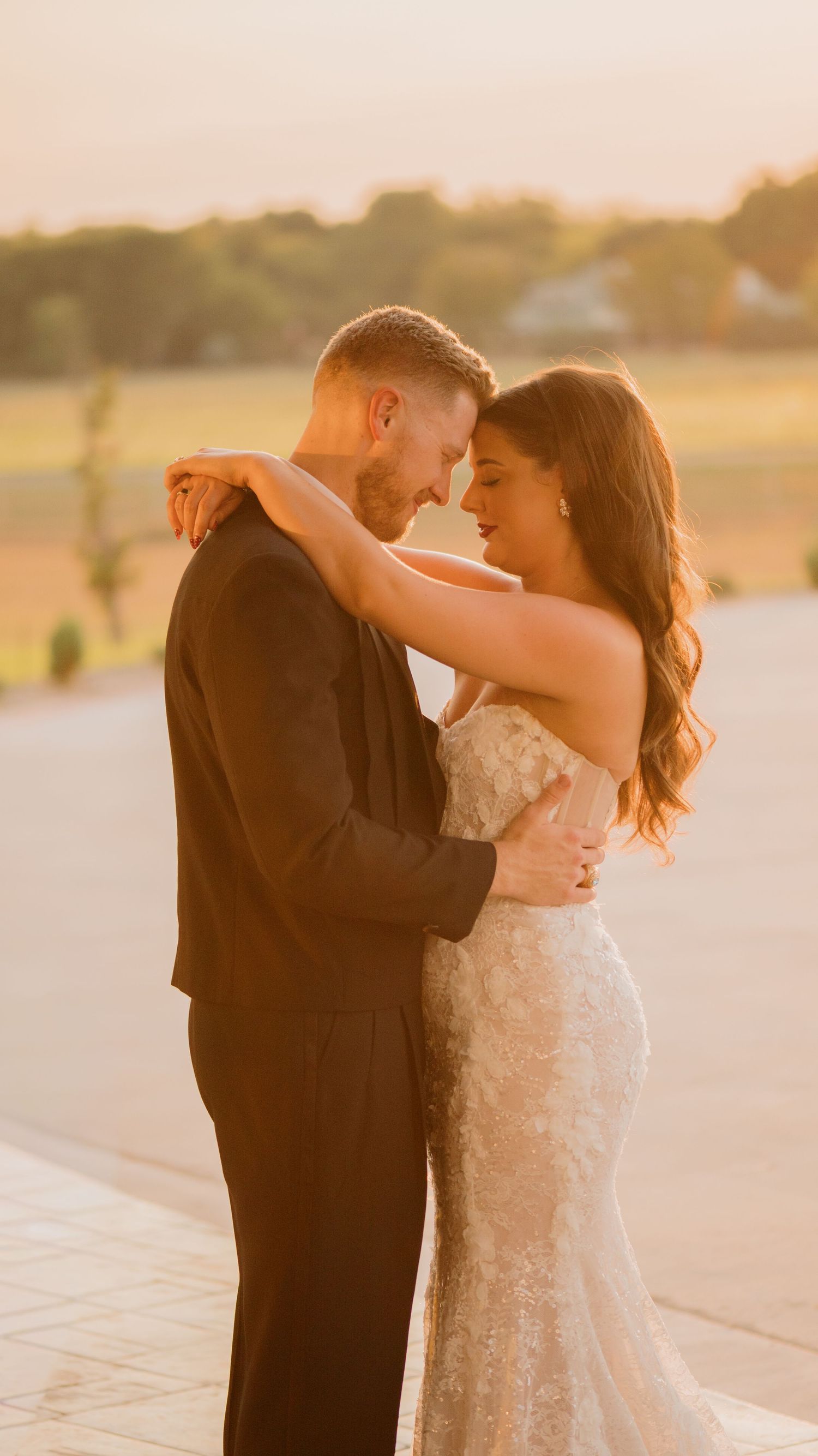 Bride and groom embrace forehead to forehead on a terrace at golden hour with rolling green fields behind them.