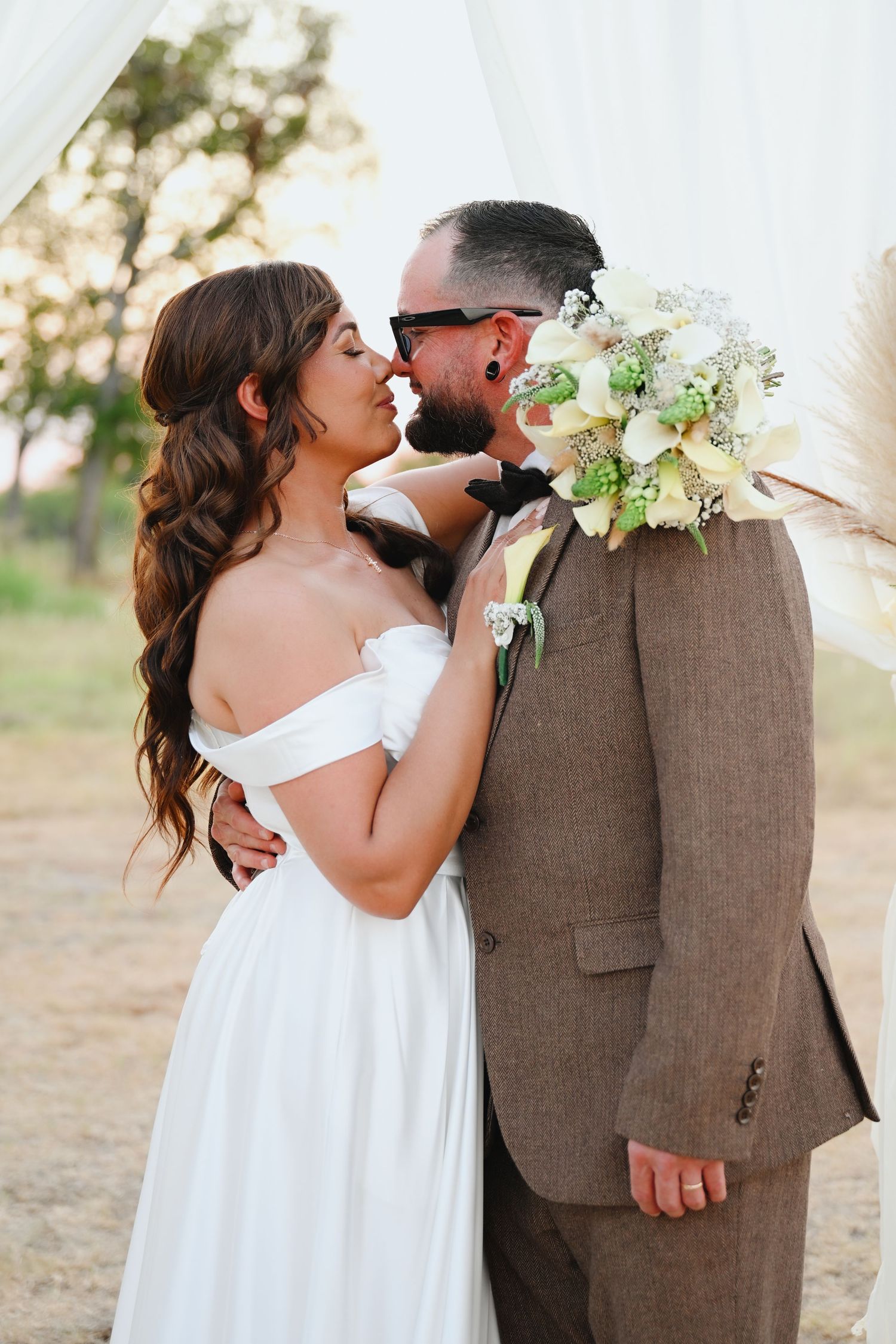 A bride in an off-the-shoulder white gown embraces her groom in a brown tweed suit and sunglasses, holding a white floral bouquet outdoors.