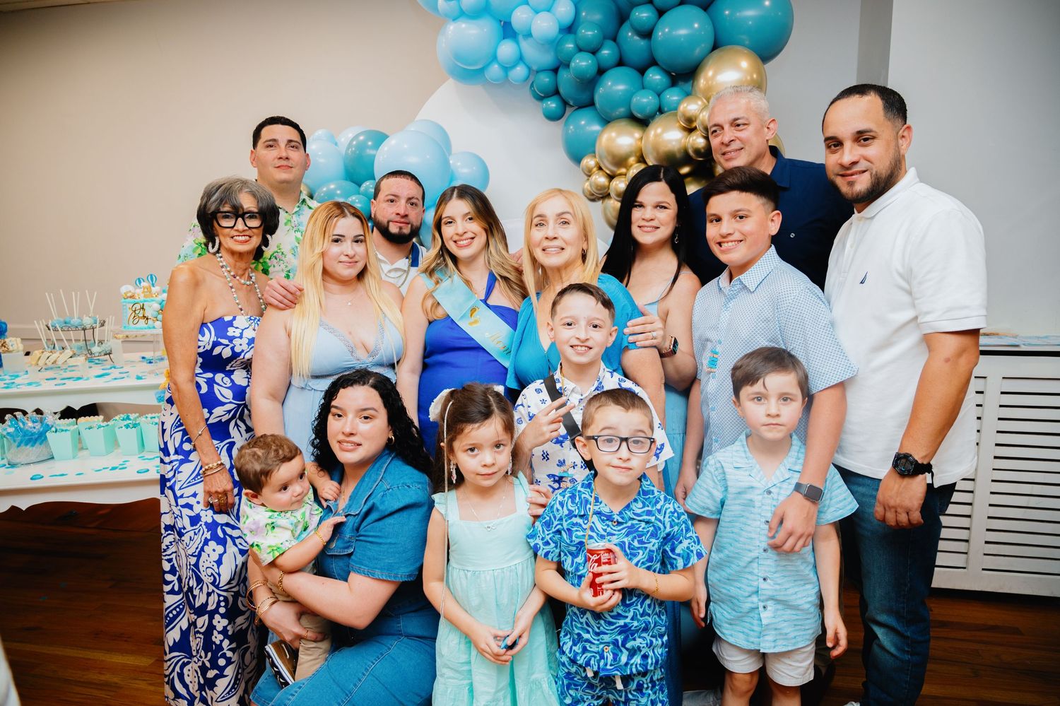 Large family and friends pose together for a group photo in front of blue and gold balloons at a celebration.