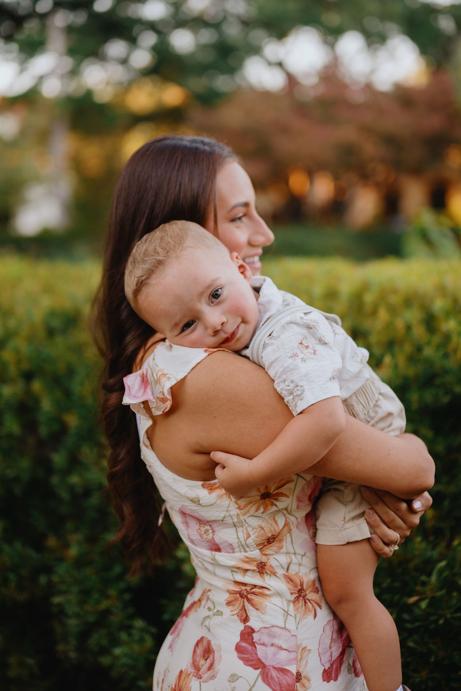 Mother in a pink floral dress hugging her smiling baby boy in a garden with lush green hedges.