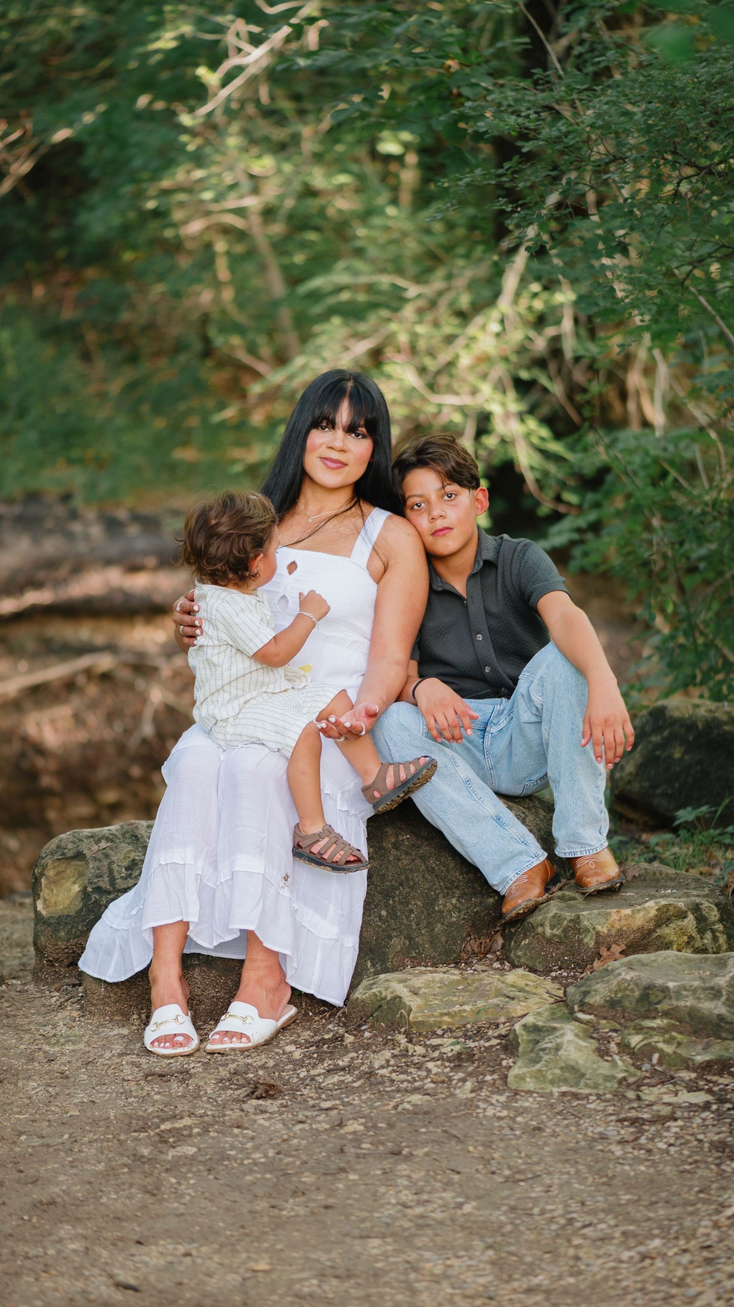 A mother in a white dress sits on rocks by a creek holding her toddler while her older son in a gray shirt sits beside her.
