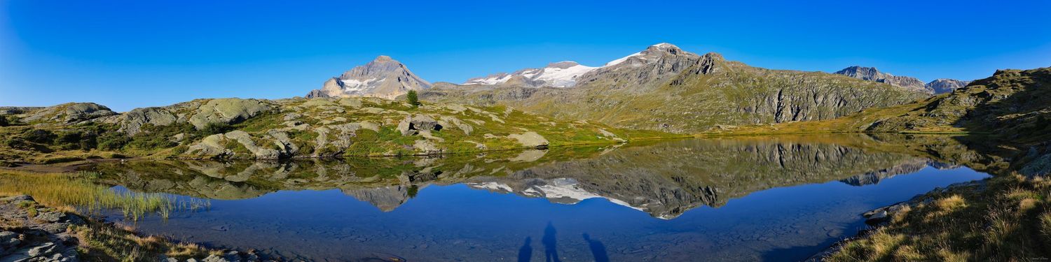 Panorama d'un lac de montagne reflétant parfaitement des sommets rocheux et enneigés sous un ciel bleu. Rives d'herbe dorée.