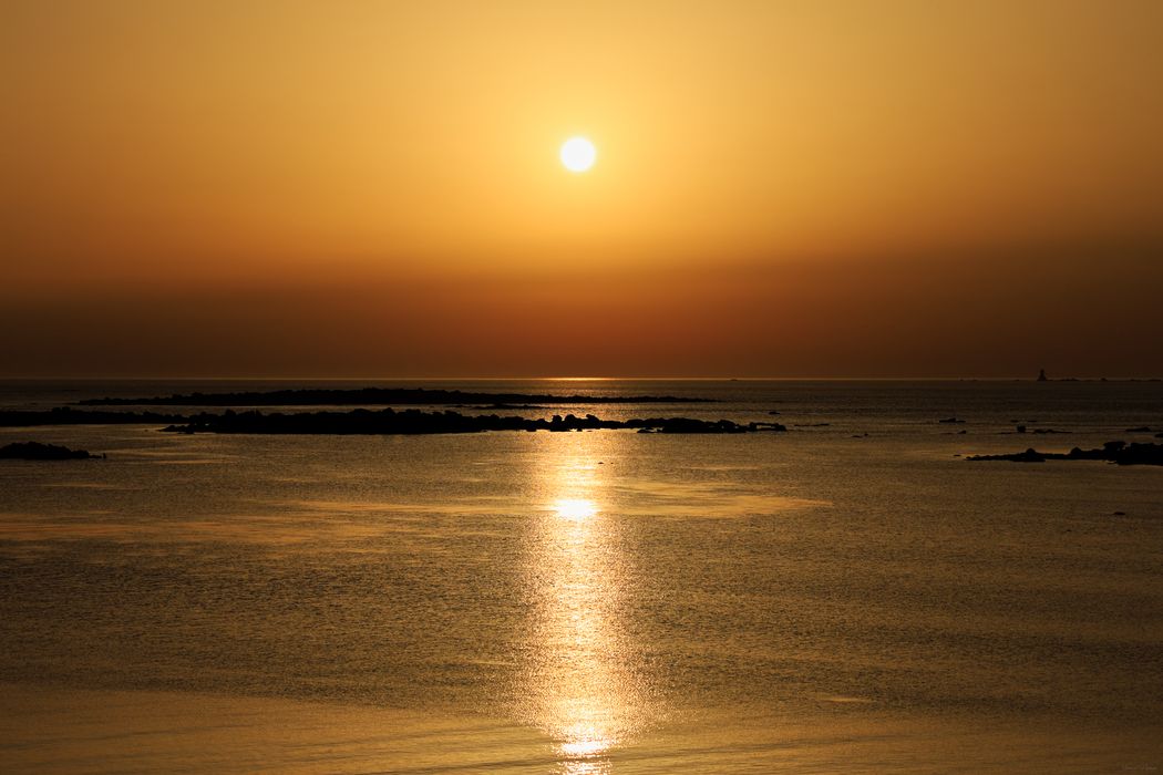 Coucher de soleil doré sur une mer calme parsemée d'îlots rocheux sombres en silhouette. Reflet du soleil sur l'eau.