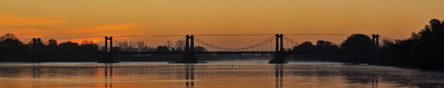Silhouette d'un pont suspendu sur un fleuve au lever du soleil. Ciel orange vif se reflétant dans l'eau calme avec une légère brume.
