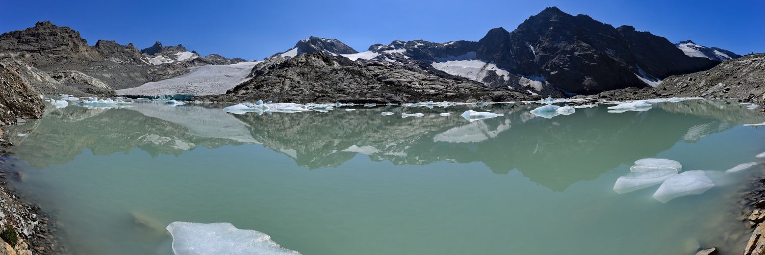 Panorama de haute montagne avec lac glaciaire turquoise. Cirque rocheux escarpé, névés et sommets sous un ciel bleu limpide.
