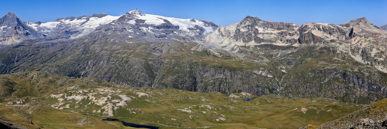 Panorama de haute montagne avec sommets rocheux et glaciers. Vallées herbeuses au premier plan parsemées de petits lacs d'altitude.