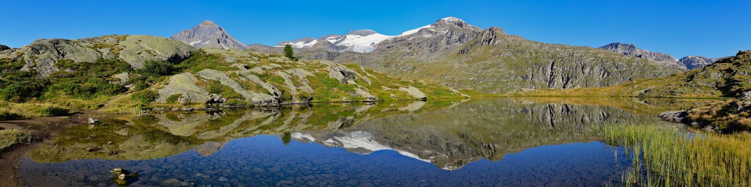 Photo panoramique d'un lac alpin limpide. Reflet parfait des sommets rocheux et enneigés dans l'eau bleue. Rives d'herbe et rochers au solei