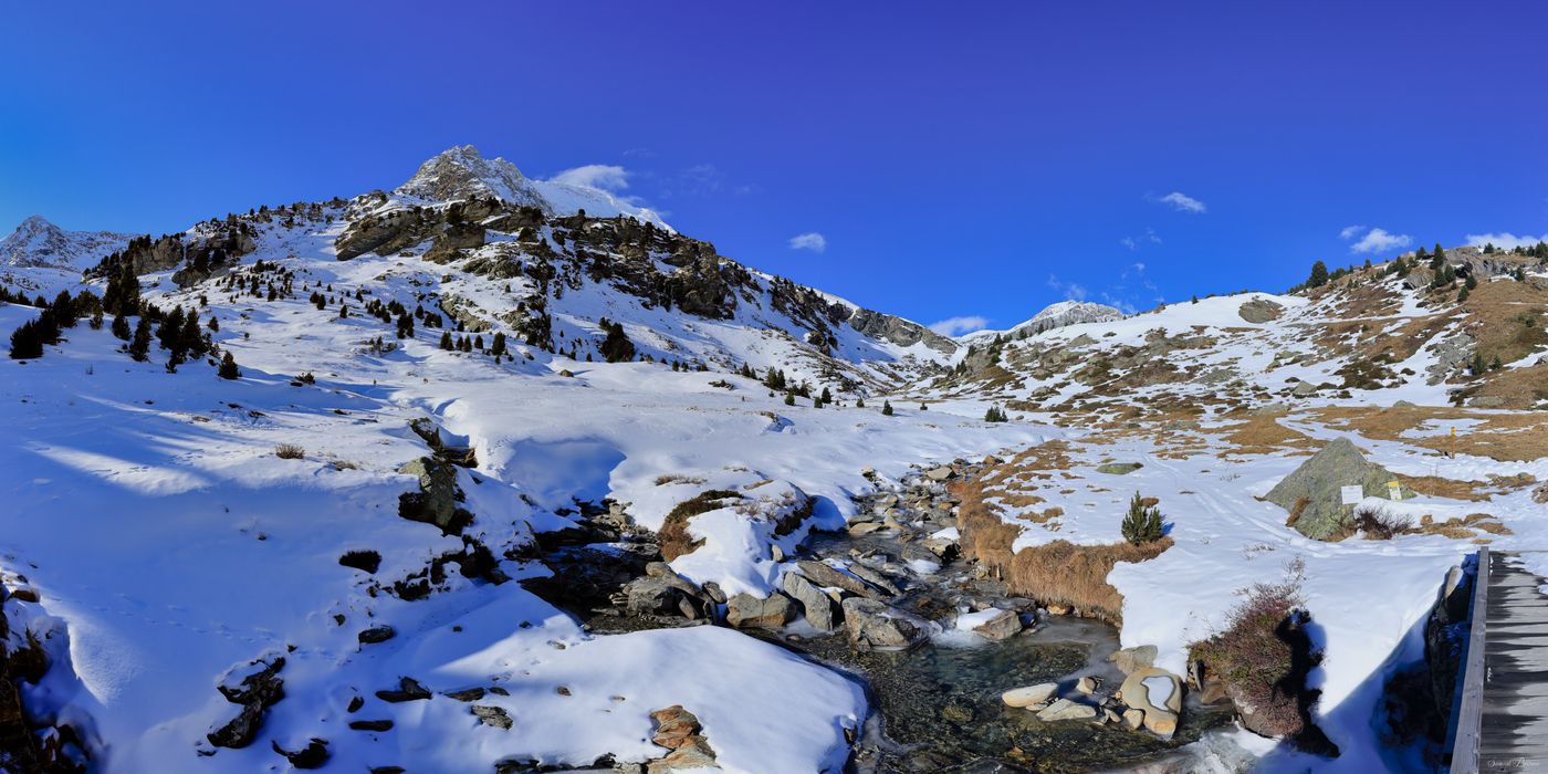 Ruisseau de montagne serpentant dans un vallon enneigé. Pont en bois au premier plan, sommets rocheux et ciel bleu profond en arrière-plan.