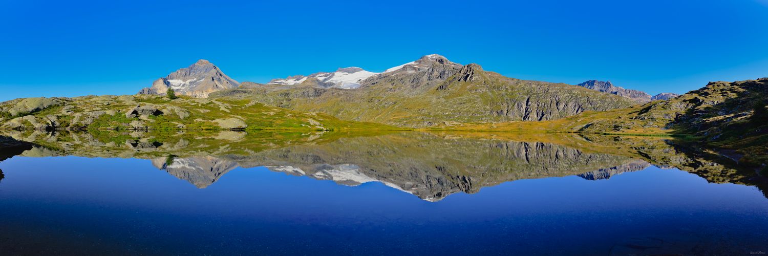 Miroir d'eau parfait d'un lac d'altitude reflétant des montagnes rocheuses et enneigées. Ciel bleu profond et rives herbeuses dorées.