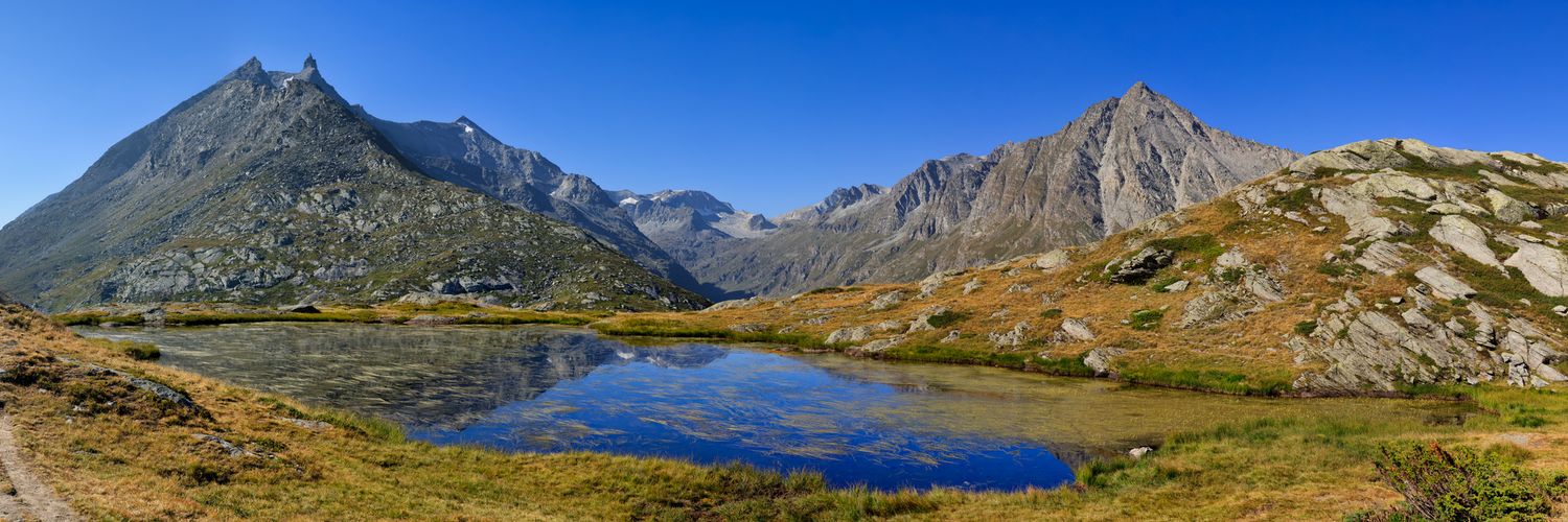 Petit lac de montagne reflétant des sommets rocheux et un ciel bleu. Vallons herbeux dorés et sentier au premier plan.