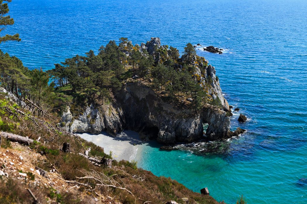 Vue aérienne plongeante sur l'îlot boisé de l'Île Vierge, sa crique cachée de sable blanc, son arche de roche et ses eaux turquoise.