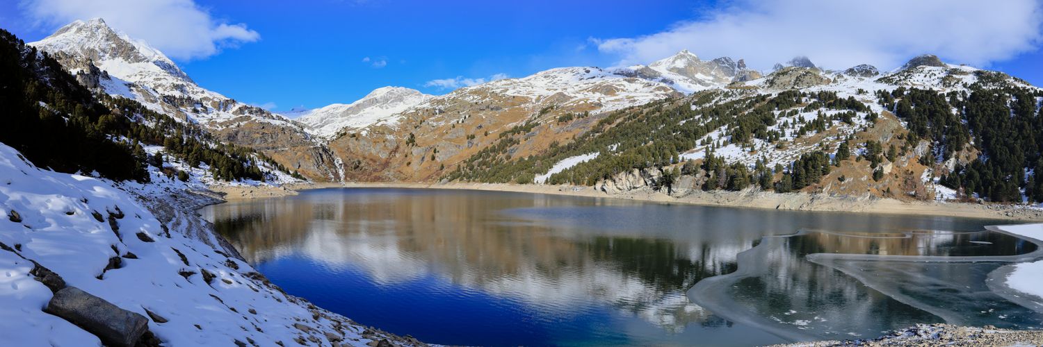 Panorama d'un lac de montagne partiellement gelé. Rives et sommets environnants recouverts de neige sous un ciel bleu parsemé de nuages.