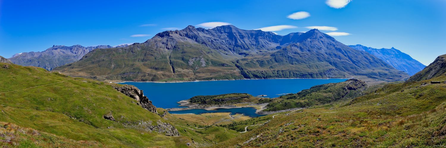 Large panorama d'un lac de montagne bleu intense entouré de pentes verdoyantes et de sommets rocheux. Ciel bleu avec quelques nuages.
