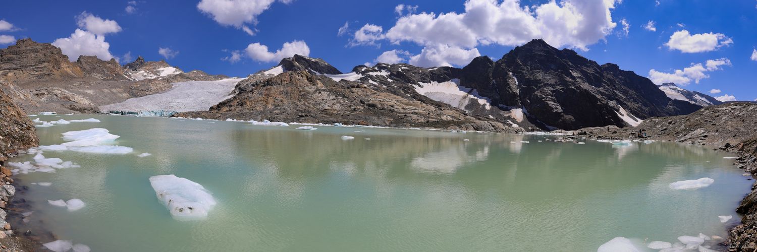 Panorama d'un lac glaciaire aux eaux vert pâle avec blocs de glace flottants. Cirque rocheux et névés sous un ciel nuageux.