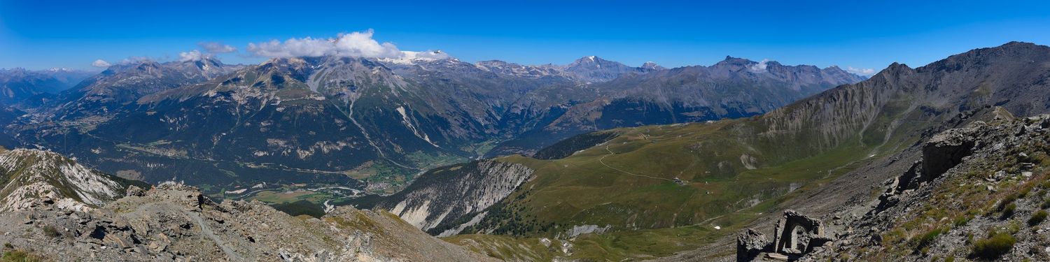 Large panorama de montagnes rocheuses et de vallées. Crêtes escarpées au premier plan, chaîne de sommets enneigés à l'horizon. Ciel clair.
