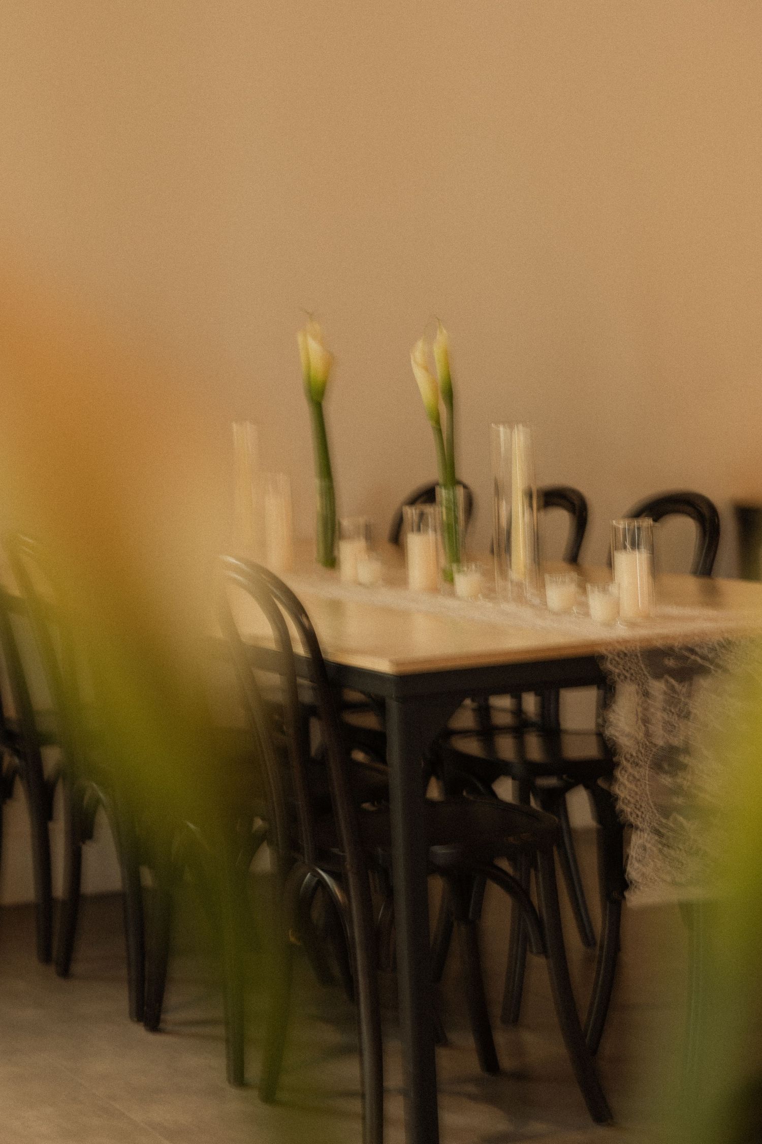 Elegant wooden dining table with black bentwood chairs, white calla lily vases and candles in a softly blurred, warm-toned setting.