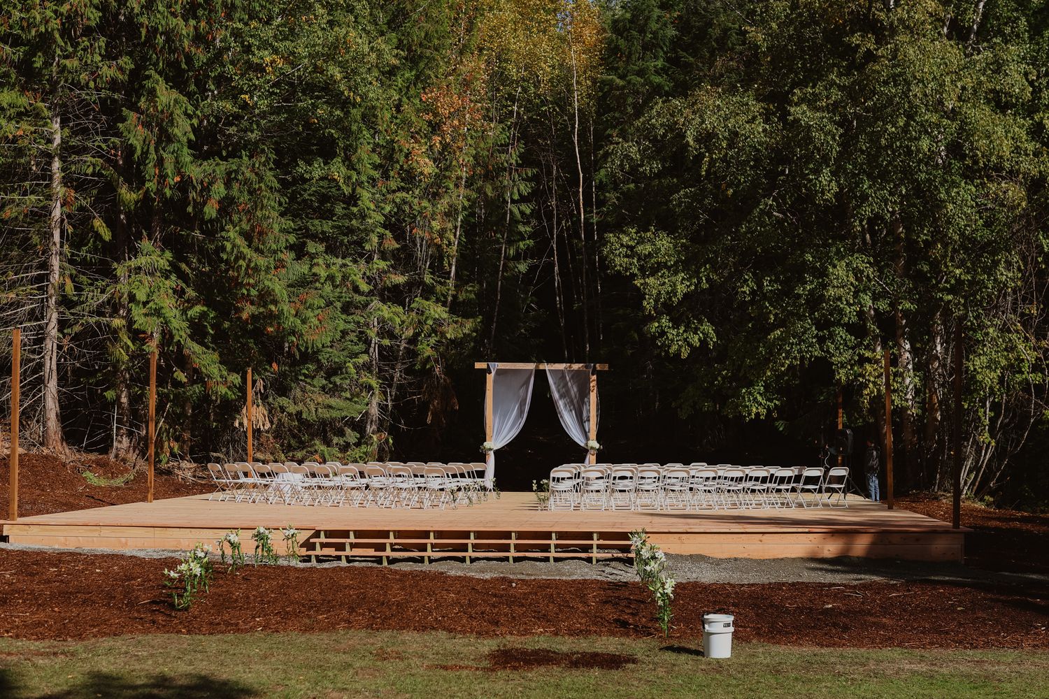 Outdoor forest wedding ceremony site with a wooden platform, draped white arbor, and rows of white chairs beneath tall evergreen trees.