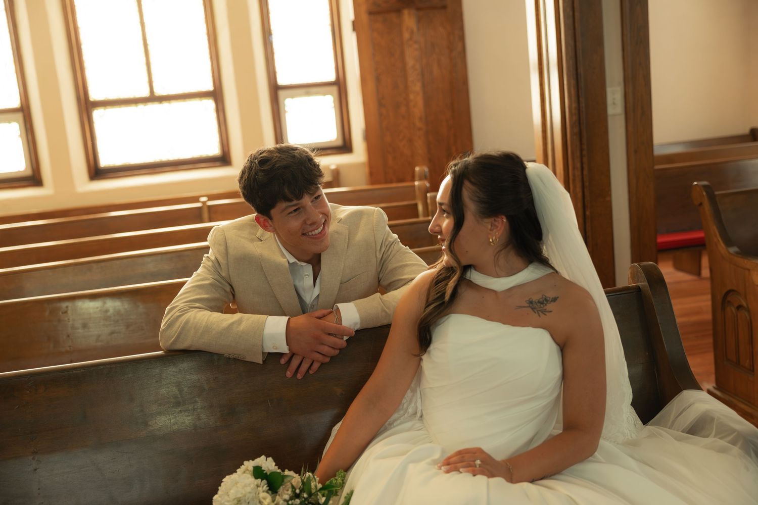 Bride in a white wedding gown smiles at a groom in a tan suit as they sit together on a wooden church pew holding a bouquet.