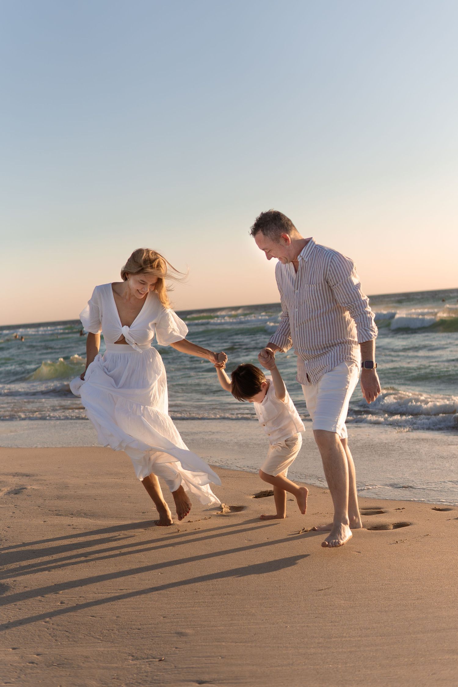 Three people in white outfits playing and jumping on a beach at sunset.