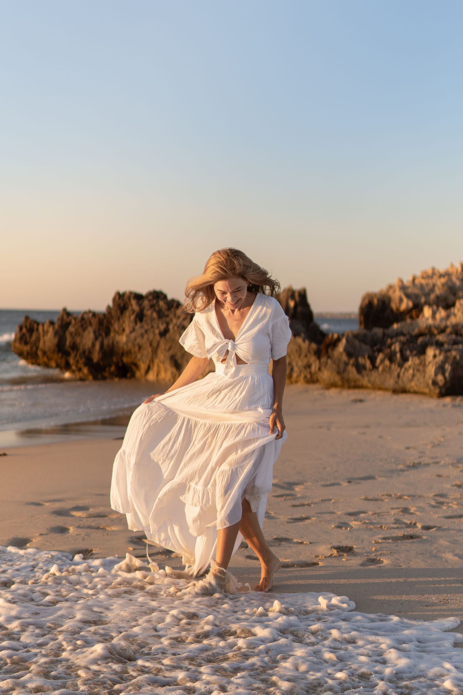 Woman in white dress dances on sandy beach with coastal rocks and ocean in background.