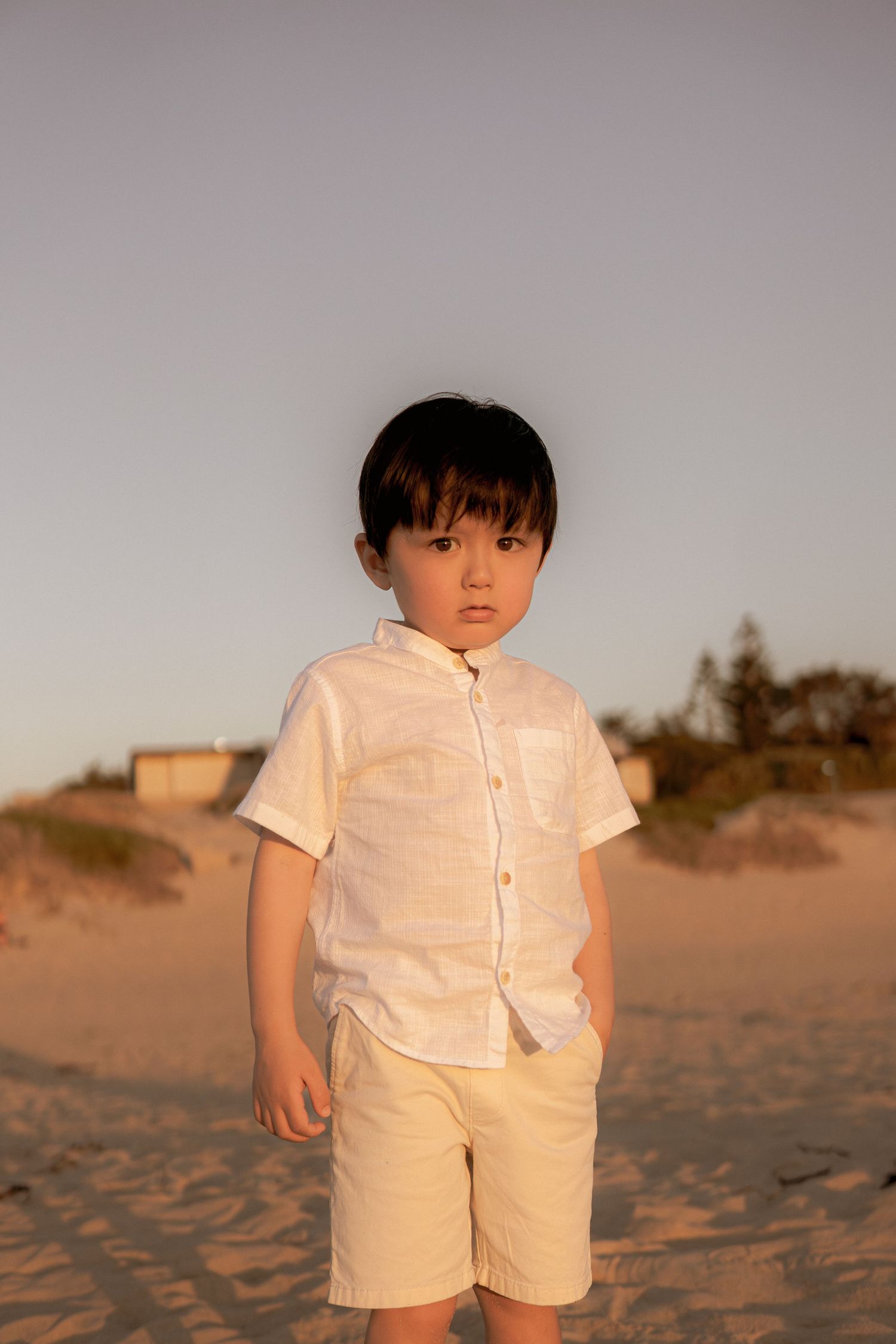 Young child in white shirt and shorts standing on sandy beach at sunset.
