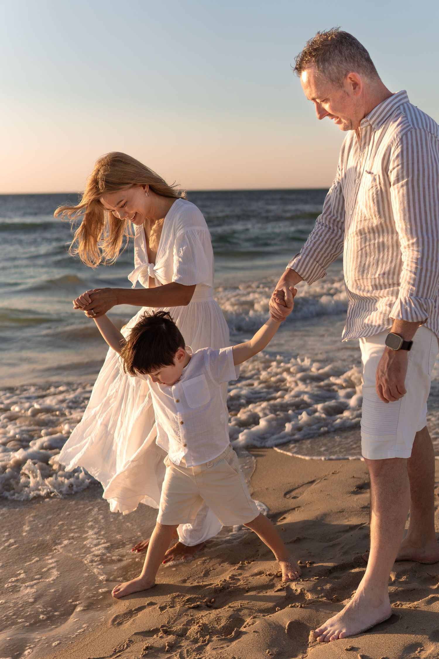 Family members in white clothing run and play on beach near ocean waves.