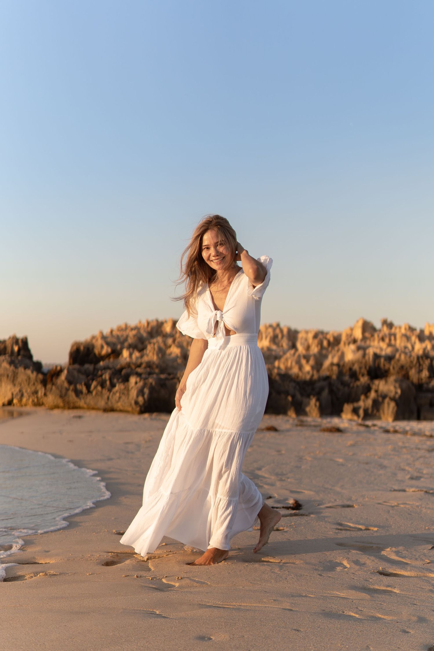Woman in flowing white dress twirls on beach with rocky coastline in background.