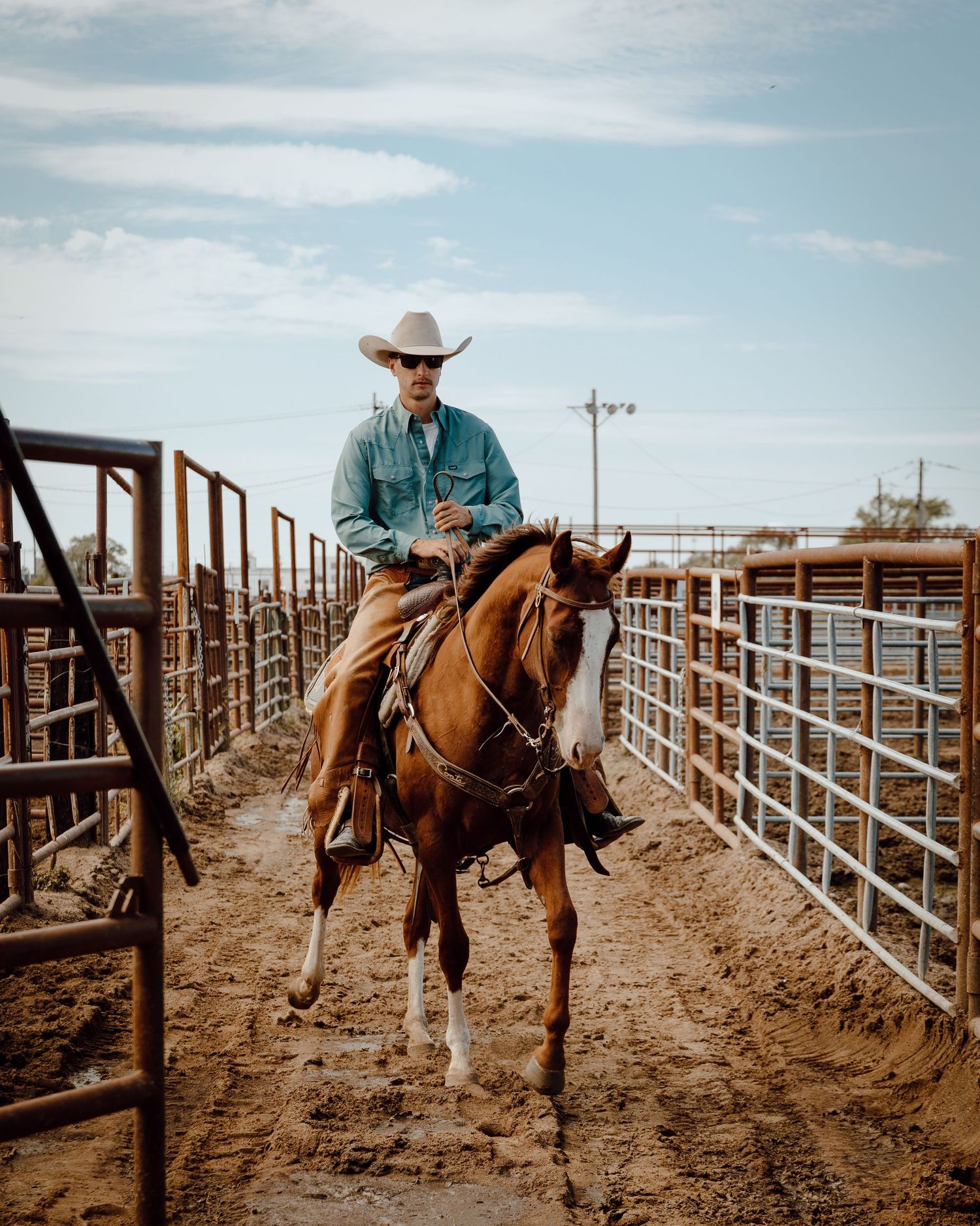 A North Platte, Nebraska lifestyle session cowboy rides horseback through a metal fenced corral under cloudy skies.