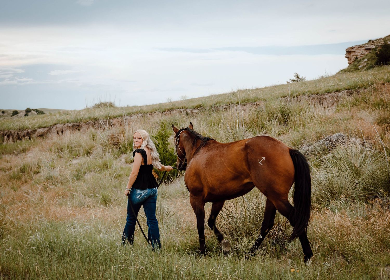 A Oshkosh, Nebraska lifestyle senior session girl leading and interacting with a brown horse in a grassy hillside pasture.