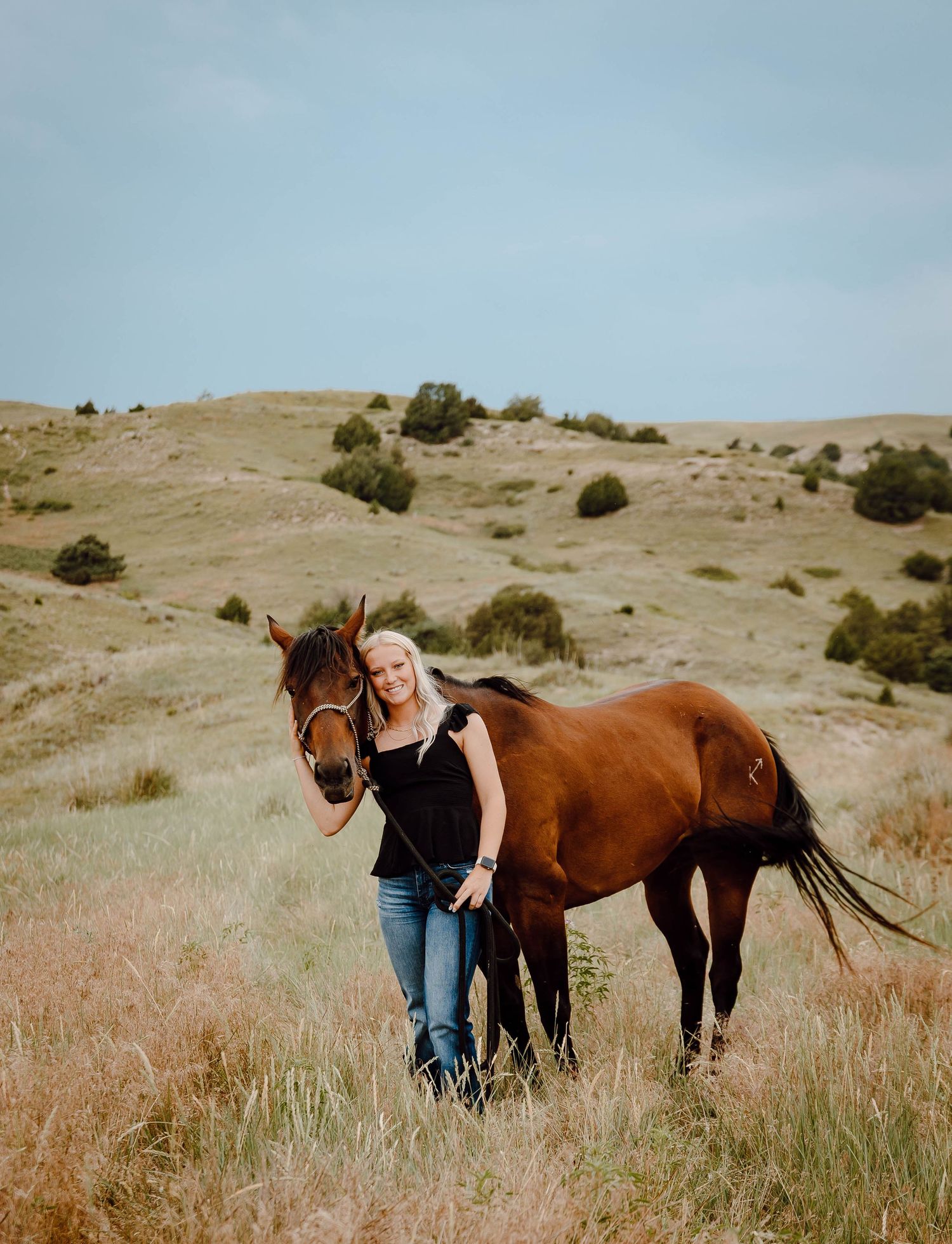 A Oshkosh, Nebraska lifestyle senior session girl standing with a brown horse in a grassy hillside field on a cloudy day.