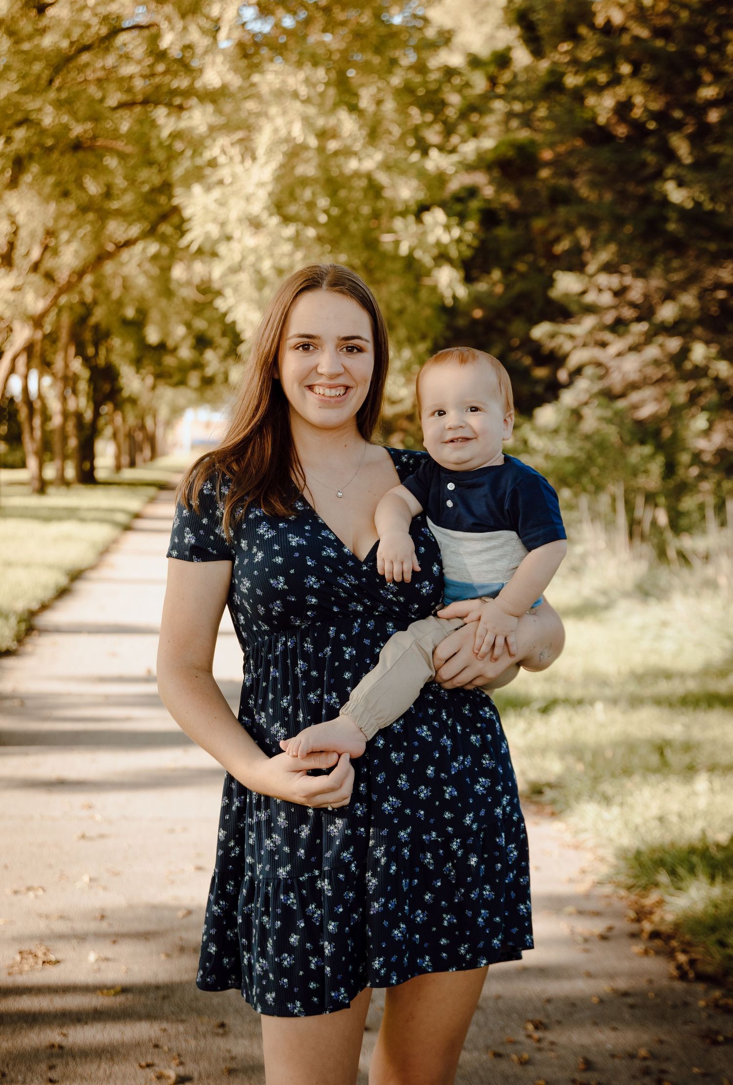 A North Platte, Nebraska lifestyle family session mom and baby stand together on a tree-lined path on a sunny day.