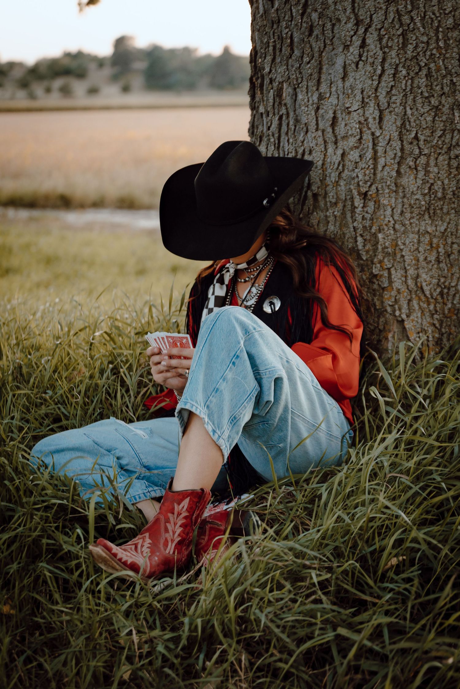 A North Platte, Nebraska lifestyle session woman in cowboy boots rests against a tree trunk in tall grass during sunset.
