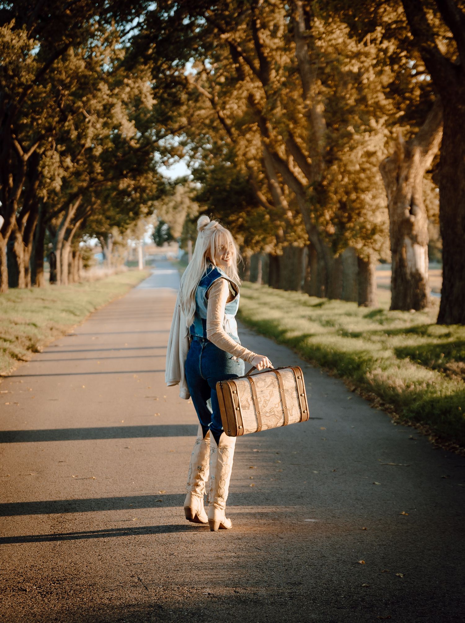 A North Platte, Nebraska lifestyle session woman walking with a suitcase in a tree-lined path with golden light filtering through branches.