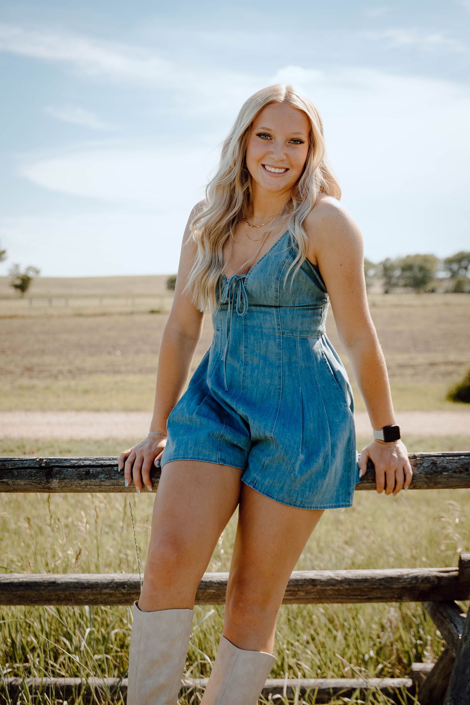 A Chappell, Nebraska lifestyle senior session girl in denim overalls poses casually against a wooden fence in a sunny field.